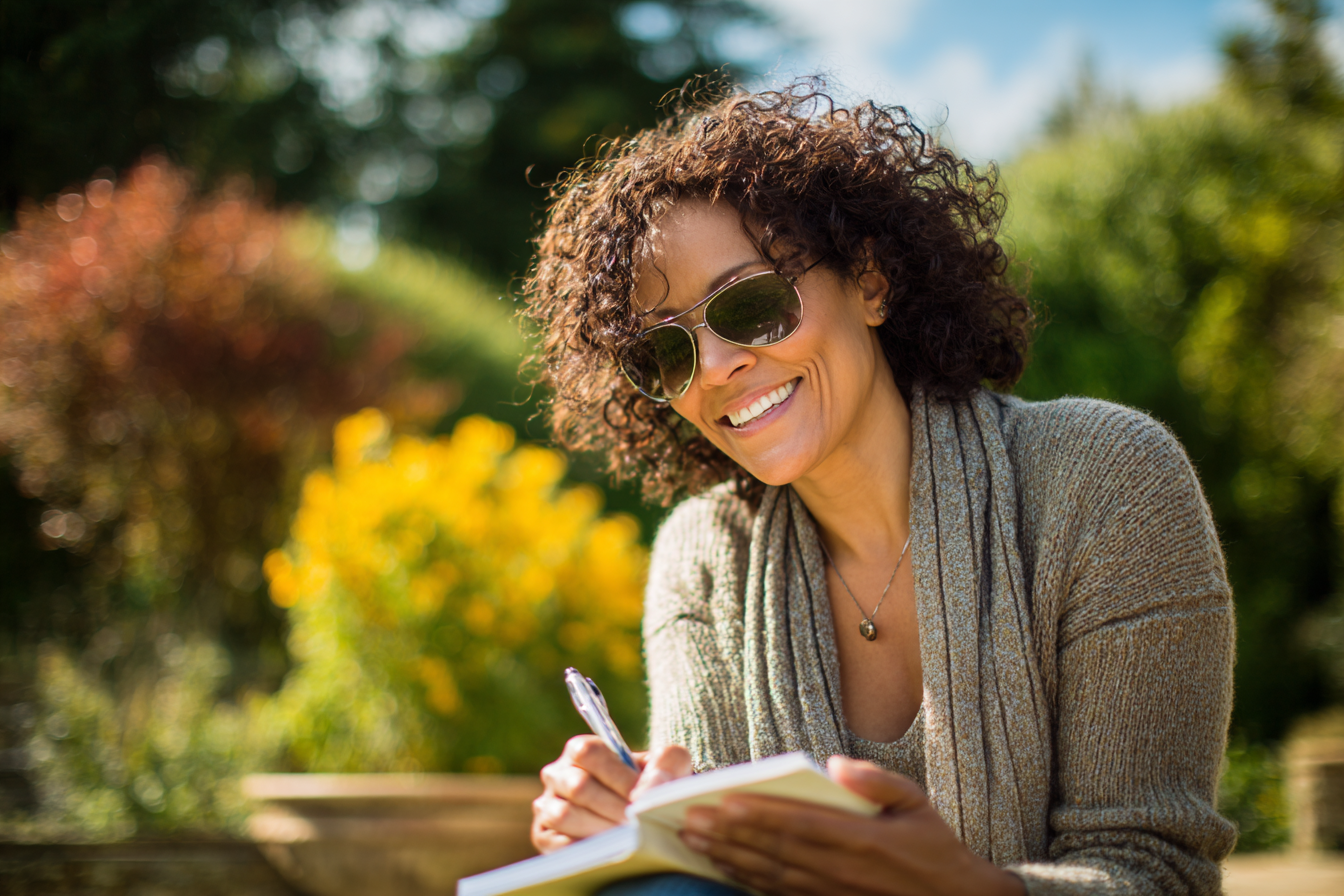 A midlife woman smiling while journaling outdoors — a visual of how to start fresh in life